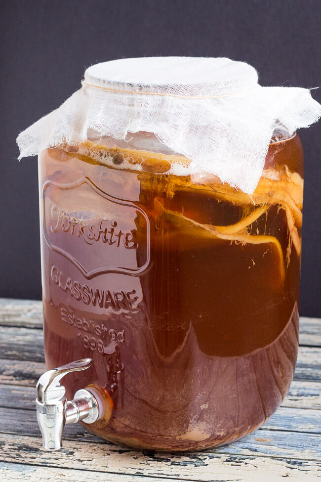 Kombucha Bryggesett 4L - Kontinuerlig Fermentering: Stor glasskrukke med klede, synlige SCOBY-lag og metallkran står på rustikk trebord-perfekt for kontinuerlig fermentering av din egen kombucha.