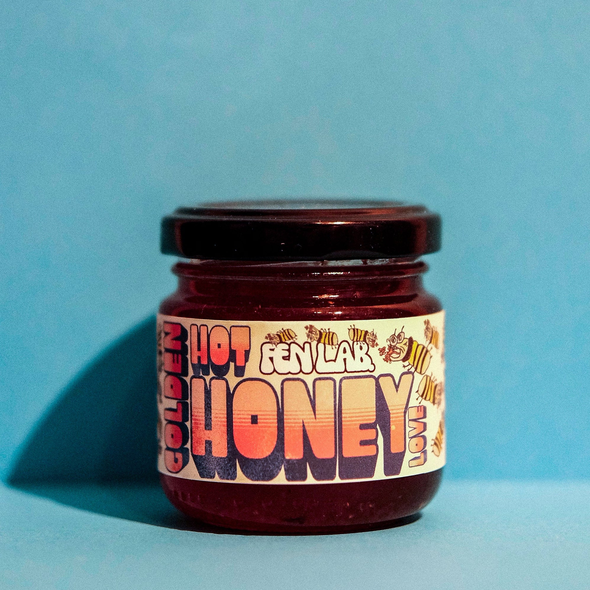 En liten glasskrukke merket Golden Hot Honey Love fra Fen Lab, med et livlig design, står på en blå bakgrunn - perfekt for å tilsette en pikant varm honningglasur til favorittmåltidene dine.