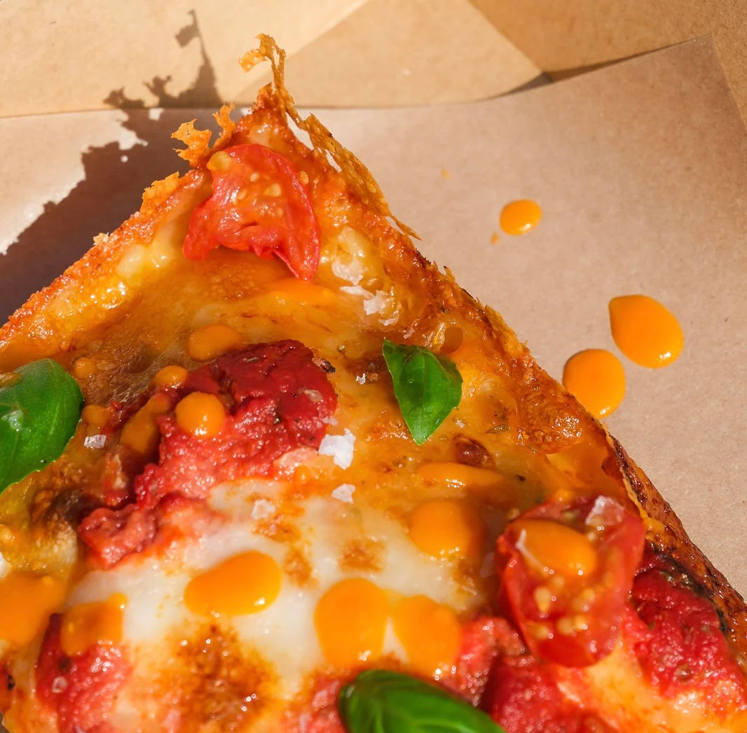 Nærbilde av en pizzaslice med smeltet ost, tomatsaus, cherrytomater, fersk basilikum og dråper av Tablesås - Den Allsidige Chilisausen | Kyrres Krutt på brunt bakepapir.