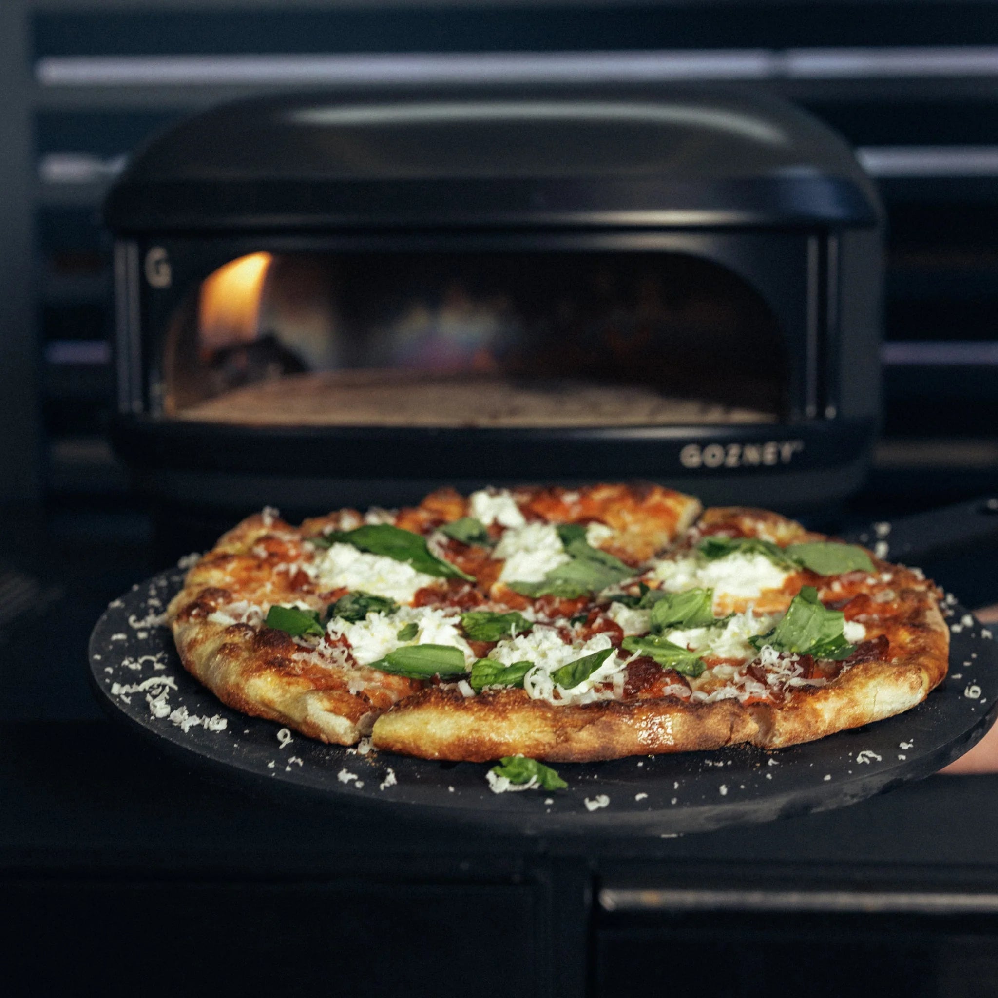 En nybakt pizza med ost, basilikum og saus holdes på et pizzaskall foran en elegant Gozney Arc Lite, Sort - Pizzaovn.