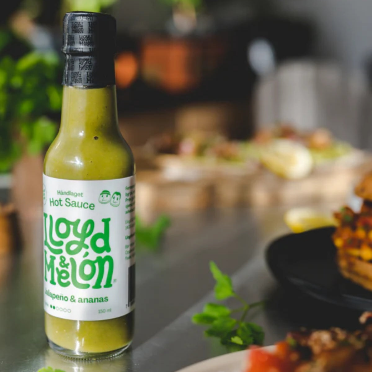 En flaske Jalapeño & Ananas Hot Sauce | Lloyd & Melón hviler på en benkeplate av metall, omgitt av uskarp mat og grøntområder - opplev den dristige, håndlagde smaken av denne unike hot sausen.