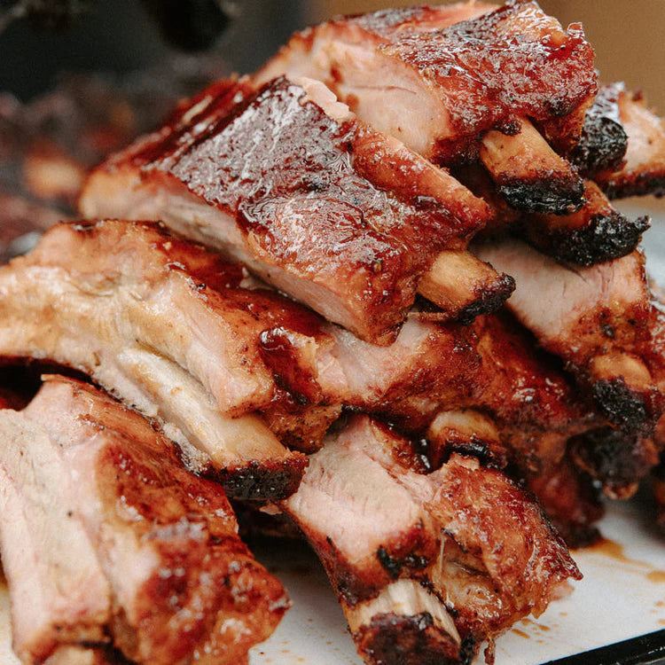 Et nærbilde av en stabel med grillede svineribber med en karamellisert, sprø overflate, som avslører saftig, mørt kjøtt og lett svidde kanter, tilberedt med Sucklebusters Honey BBQ Glaze & Finishing Sauce 355 ml for å gi dem en uimotståelig glans.