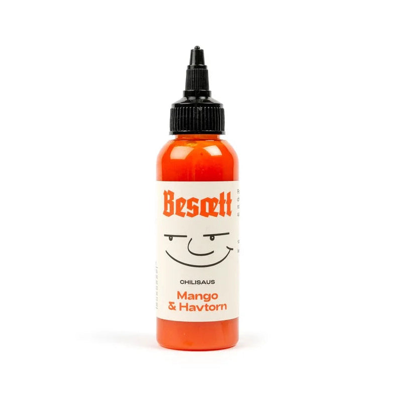 En flaske «Besætt Mango & Havtorn Fermentert Chilisaus», med en illustrasjon av et smilende ansikt på forsiden og en Carolina Reaper-chili, isolert på hvit bakgrunn.
