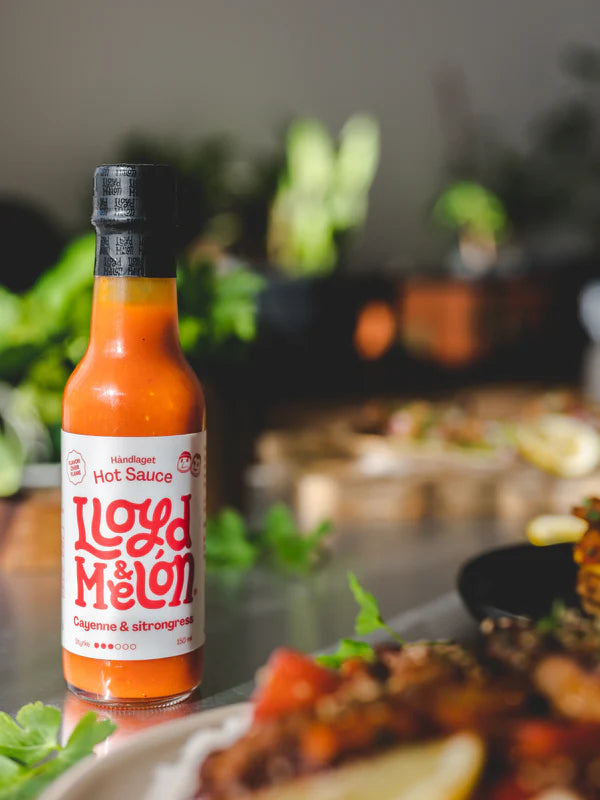 En flaske Cayenne & Sitrongress Hot Sauce | Lloyd & Melón, laget med naturlig fermentering, står på et bord ved siden av tallerkener med mat, med frodig grønt i bakgrunnen.