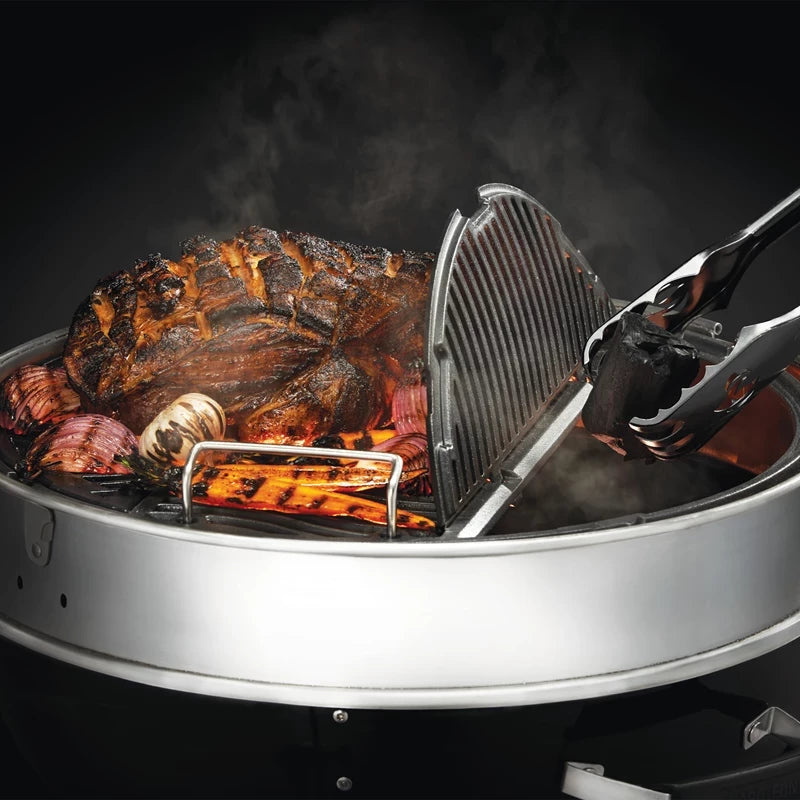 Nærbilde av en rund Napoleon PRO22 Kulegrill 57 cm med forkullet kjøtt, løk og hvitløk som syder mens tangen løfter risten; røyken stiger opp - ideelt for å mestre smaker på denne førsteklasses grillen.