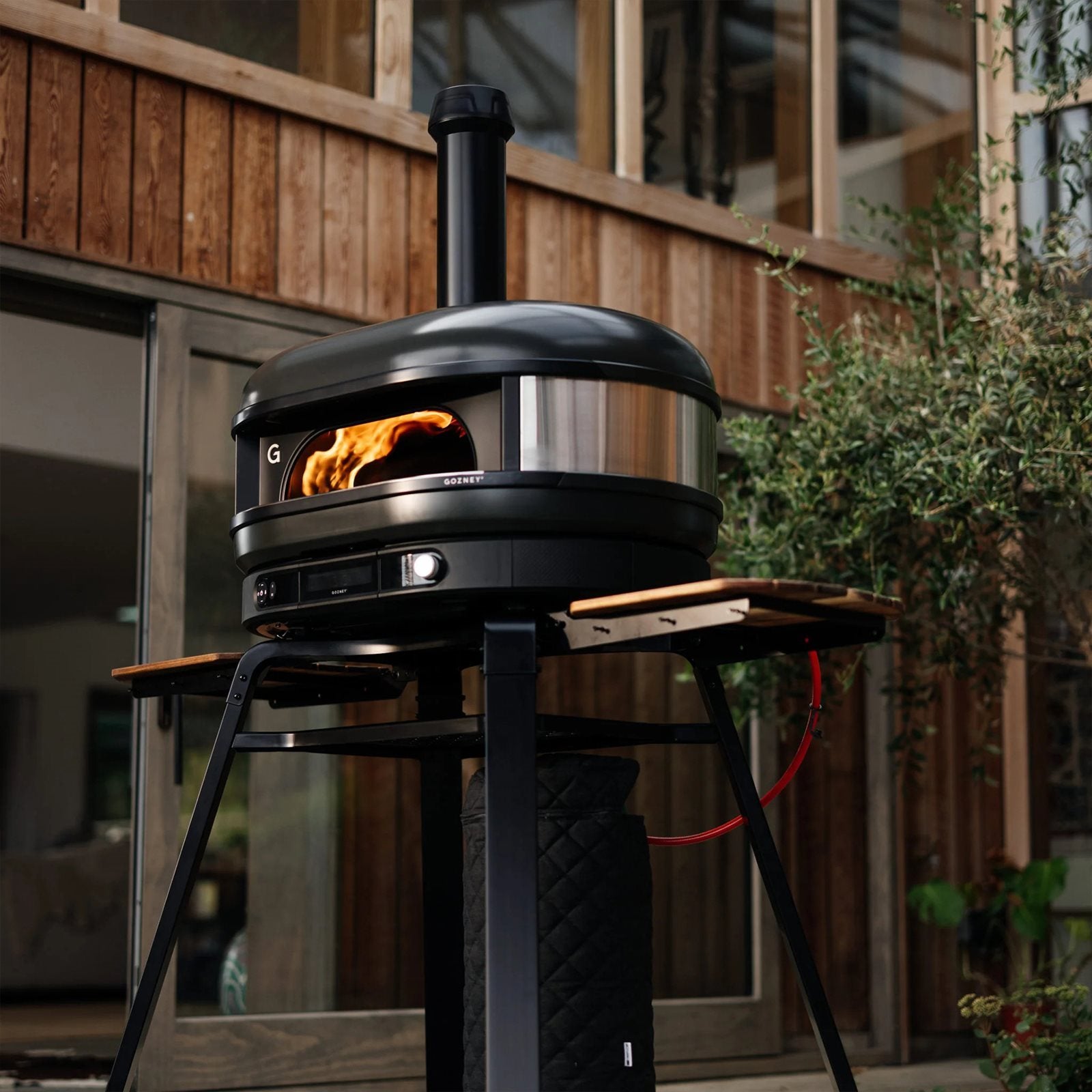En Gozney Dome Gen 2 - Off Black - Multi Fuel Pizzaovn med synlig flamme er satt opp på et stativ foran et trehus med store vinduer.