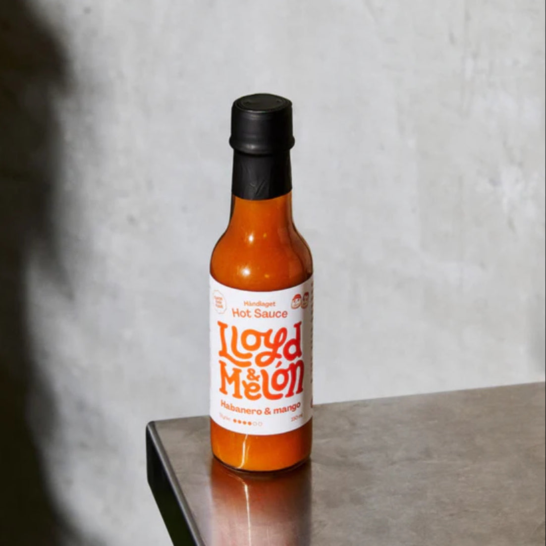 En flaske Lloyd & Melón Habanero & Mango Hot Sauce står på en metallisk overflate mot en grå vegg.