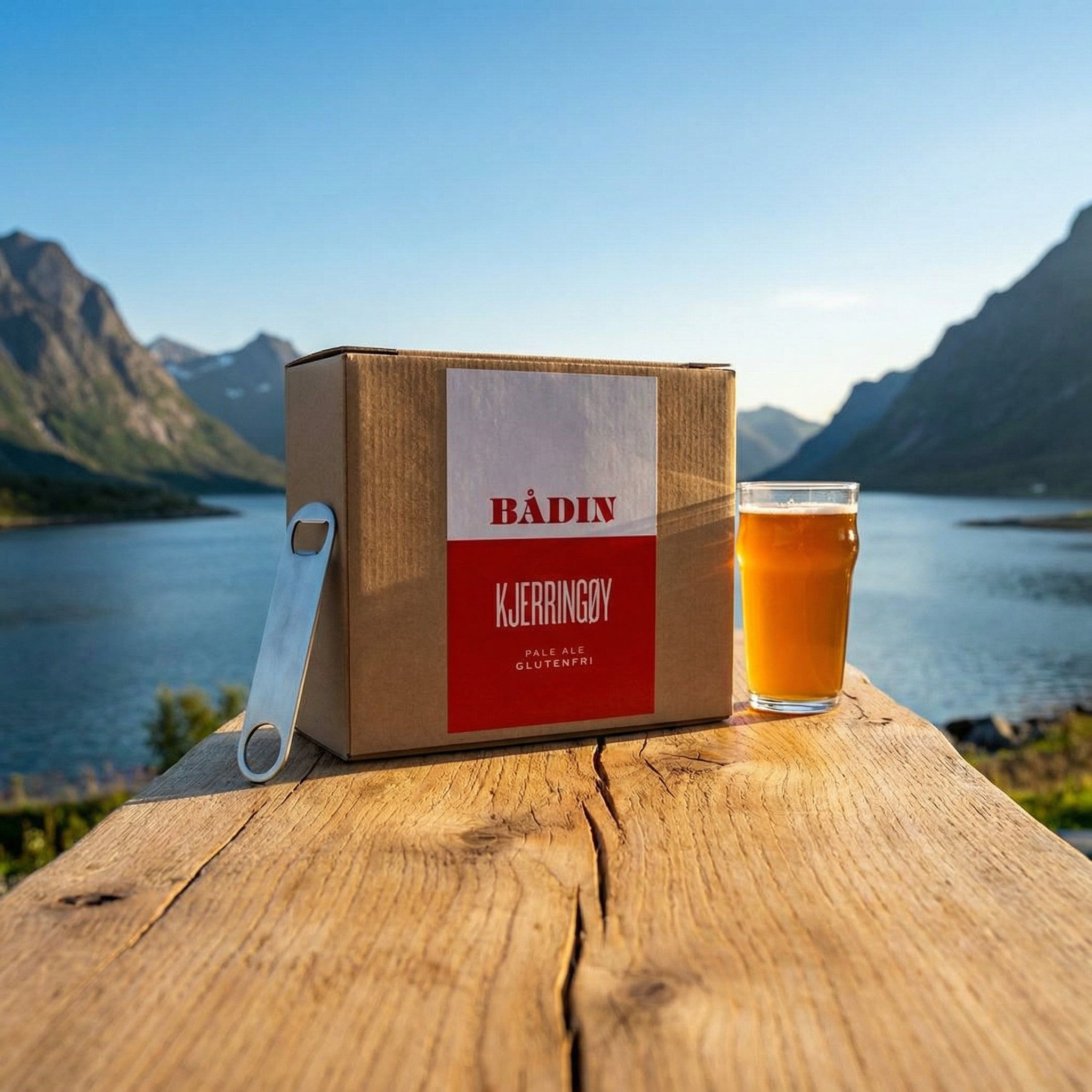 En eske merket Bådin Kjerringøy Pale Ale - allgrain ølsett og et glass Kjerringøy Pale Ale står på et trebord utendørs, med fjell, en innsjø og blå himmel i bakgrunnen.