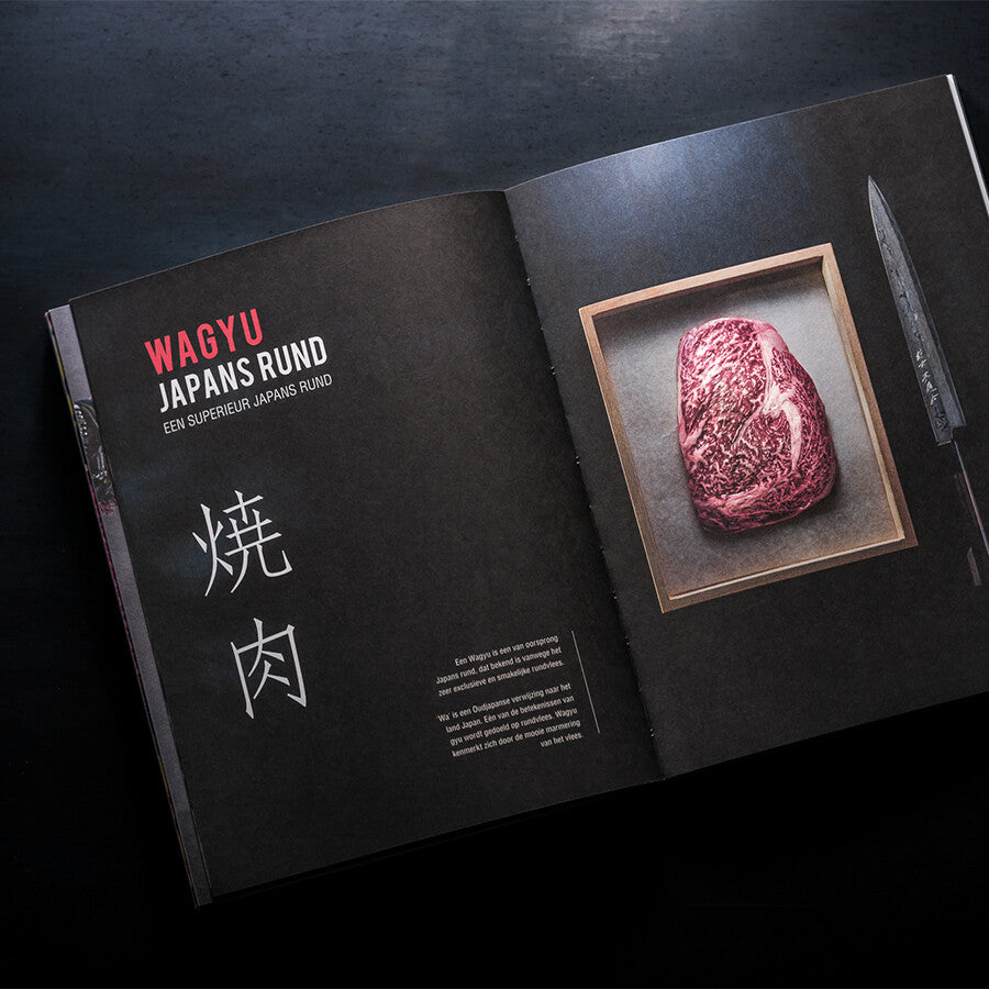 Åpent magasin med kokeboken Japanese Grill YAKINIKU, Shichirin Kokebok, som viser et marmorert Wagyu-biffstykke skåret i et trebrett, en kniv, japanske tegn og nederlandsk tekst om Yakiniku og tradisjonell japansk grillkunst.