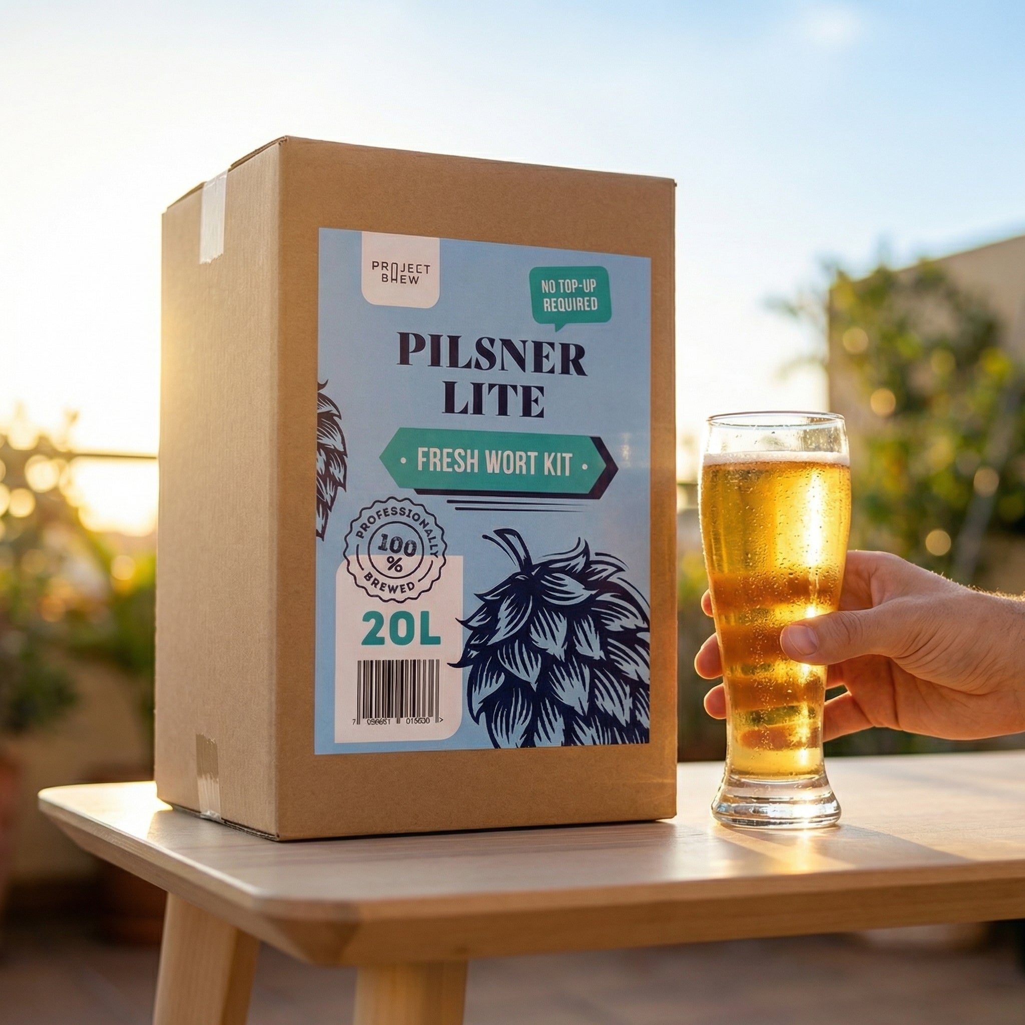 En eske merket Pilsner Lite - 20L Fresh Wort Kit står på et bord ved siden av en hånd som holder et glass lavkarboøl, mens sollyset strømmer inn i bakgrunnen.