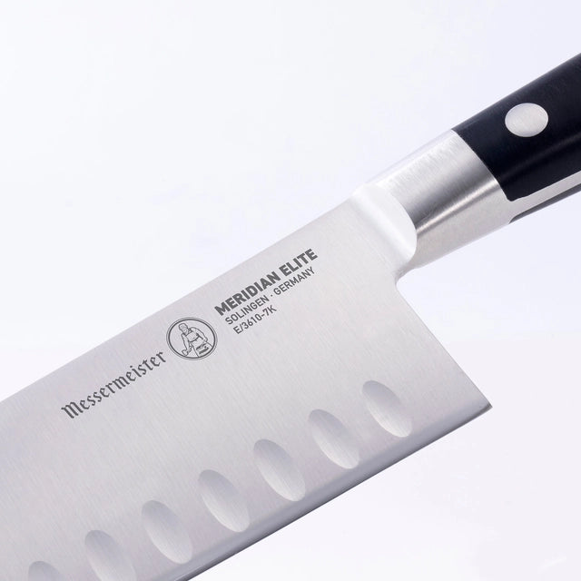 Et nærbilde av Messermeister Meridian Elite 18 cm Kullenschliff Santoku japansk kokkekniv, med et elegant svart håndtak og karakteristisk gravering på bladet, som utstråler presisjon og eleganse på høyde med enhver japansk kokkekniv.