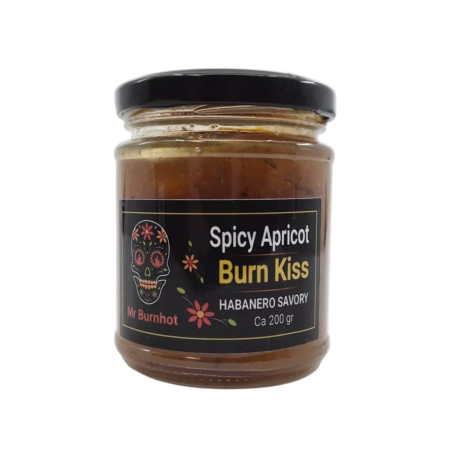 En glasskrukke på 200 gram med «Burn Kiss Spicy Apricot» fra Mr Burnhot har en fargerik illustrasjon av et hodeskalle med blomsterdetaljer på den svarte etiketten – denne håndlagde syltetøyen er mer enn bare syltetøy; den kombinerer på kunstnerisk vis krydret aprikos og brennende habanero for en uforglemmelig smaksopplevelse.