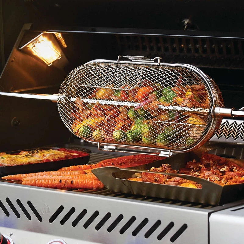 Grønnsaker og grillspyd tilberedes i en Napoleon Rotisserikurv, en roterende nettingkurv av rustfritt stål, på en grill, med brett med grilltilbehør som gulrøtter og innbakte retter under.