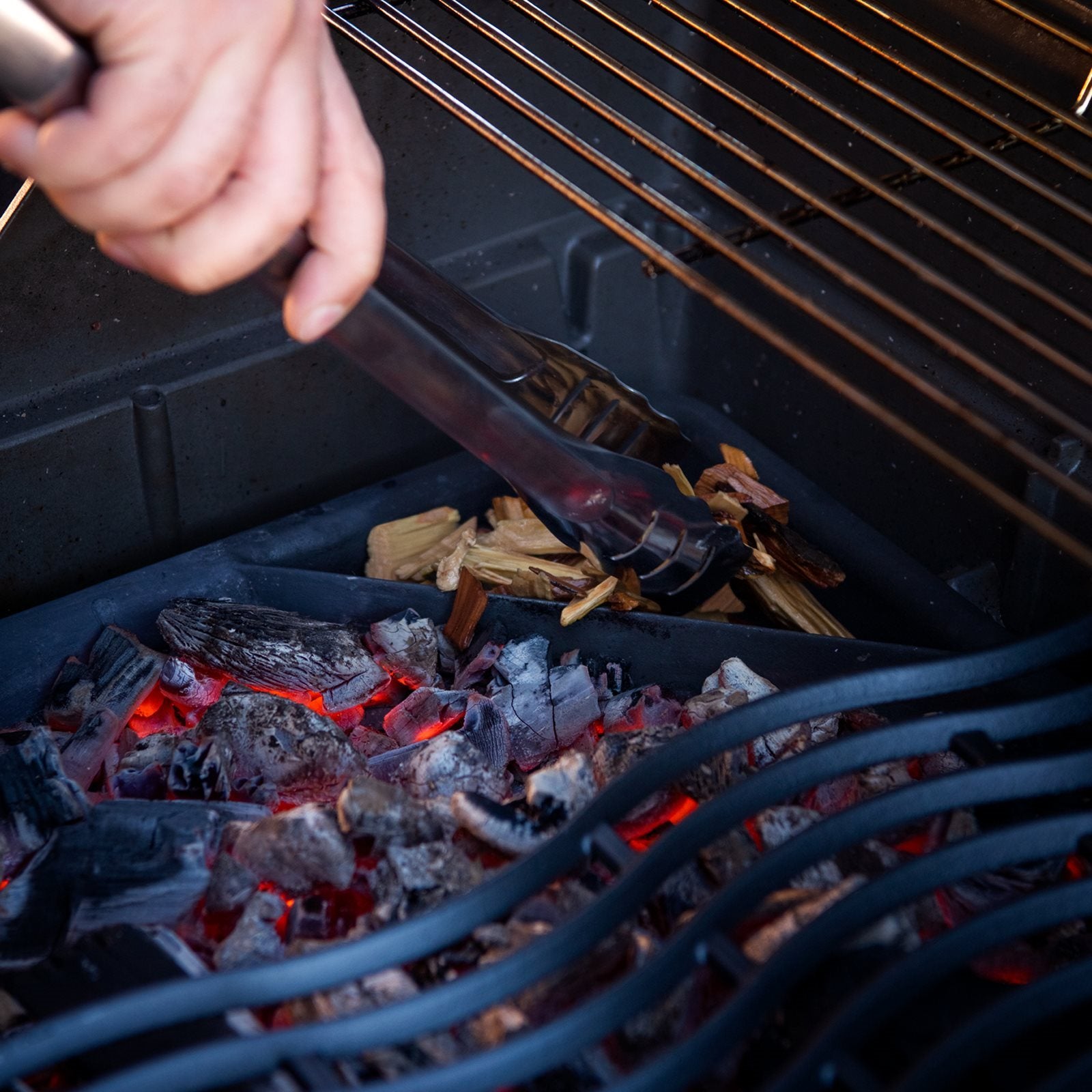 En hånd bruker en metalltang til å plassere treflis på varmt trekull inne i et Napoleon Grillkullrist - kull- og røykebrett, noe som gir grillen en røkt smak.
