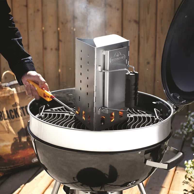 En person bruker en Napoleon sammenleggbar grillstarter til å tenne briketter på en rund kullgrill med åpent lokk, utendørs i nærheten av et tregjerde.