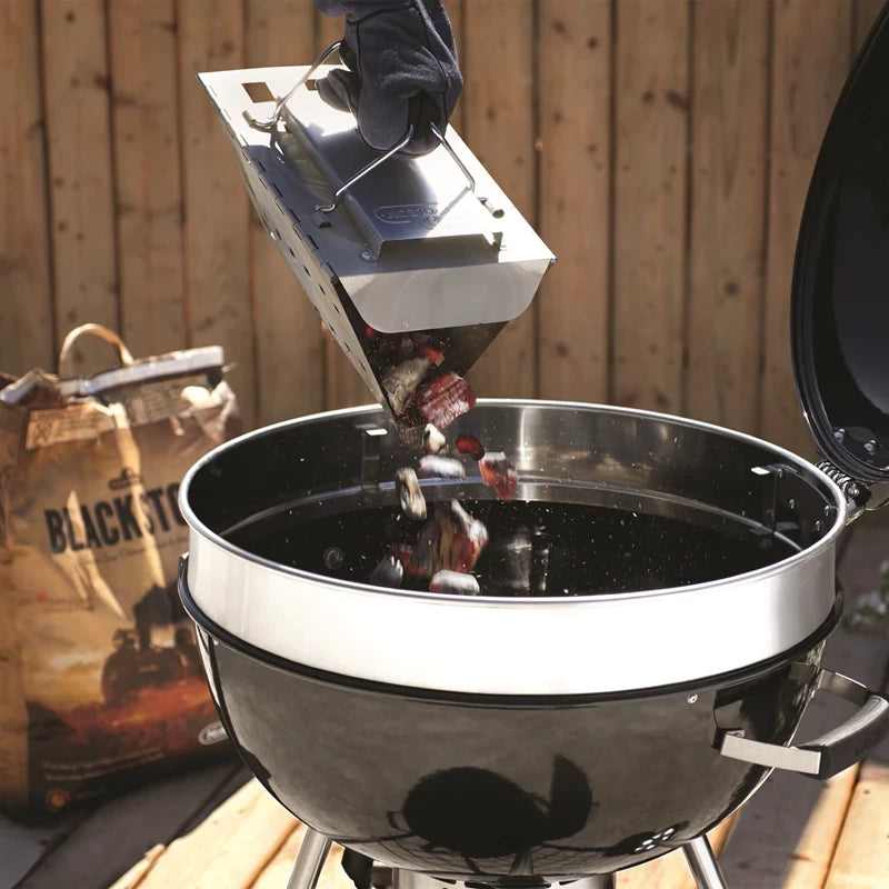 En hanskekledd hånd heller varmt kull fra en Napoleon sammenleggbar grillstarter over i en rund, svart kullgrill utendørs, med en pose briketter i nærheten.