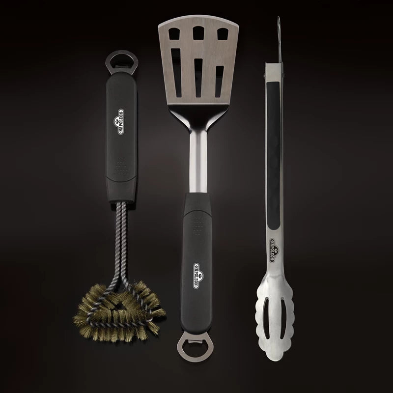 Napoleon-grillsettet med grilltang, palett og børste er avbildet vertikalt mot en mørk bakgrunn, og består av en grillbørste, en spaltespatel (palett) og en grilltang – alle med elegante, svarte håndtak.