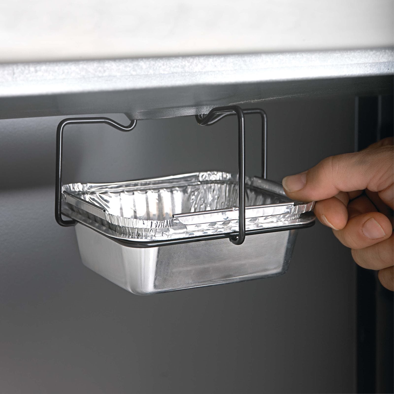 En hånd skyver en Napoleon Aluminiumsformer, stekefett oppsamler 5stk inn i en svart metallholder under en flate, klar for effektiv grillrensing.