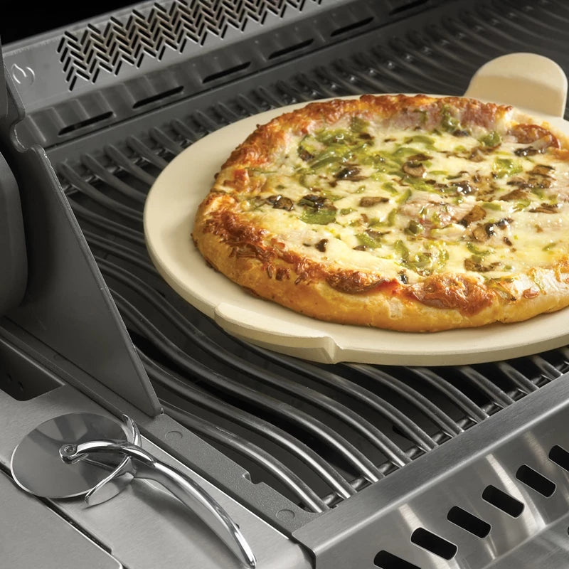 En ferdigstekt pizza med sopp og grønn paprika ligger på Napoleon PRO Pizzastein med pizzahjul inne i en grill, noe som lover ekstra sprø pizzabunn. Det medfølgende pizzahjulet hviler like ved på grillristen.