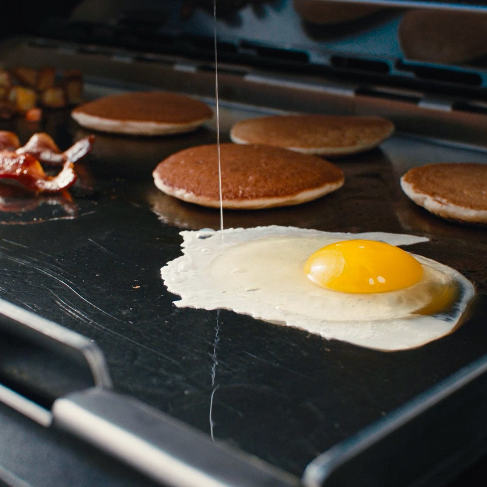 Nærbilde av et stekt egg, pannekaker og bacon som syder på en Napoleon Plancha til 425 i rustfritt stål, med en tynn strøm av væske som helles på egget.