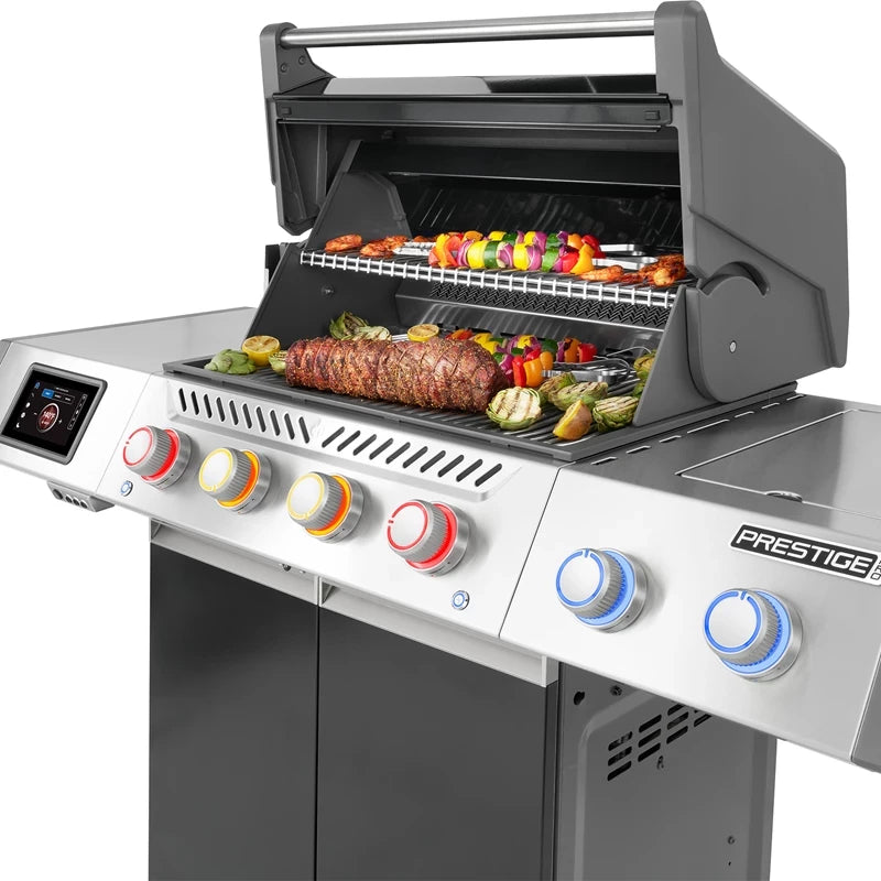 Napoleon Prestige PRO™ 500 Connected, rustfritt stål/svart (bestillingsvare), har et delvis åpent lokk som viser grillspyd med grønnsaker og kjøtt inni; betjeningsknappene og den kraftige infrarøde SIZZLE ZONE er synlige på forsiden.