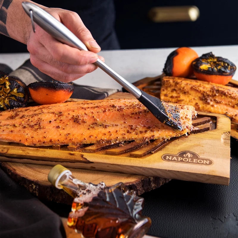 Noen bruker en Napoleon PRO-silikonpensel til å pensle marinade på en laksefilet som ligger på en Napoleon-treplank, med grillede appelsiner og lønnesirup ved siden av.