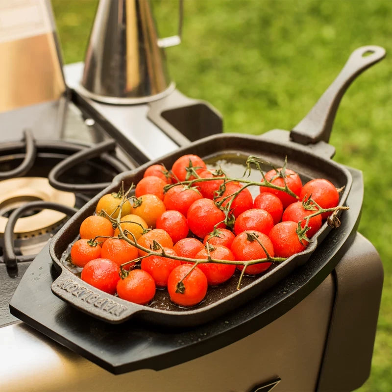 En Napoleon PRO Sizzle-panne i støpejern med vintomater står på en grill ved siden av en gassbrenner utendørs, med grønt gress i bakgrunnen.