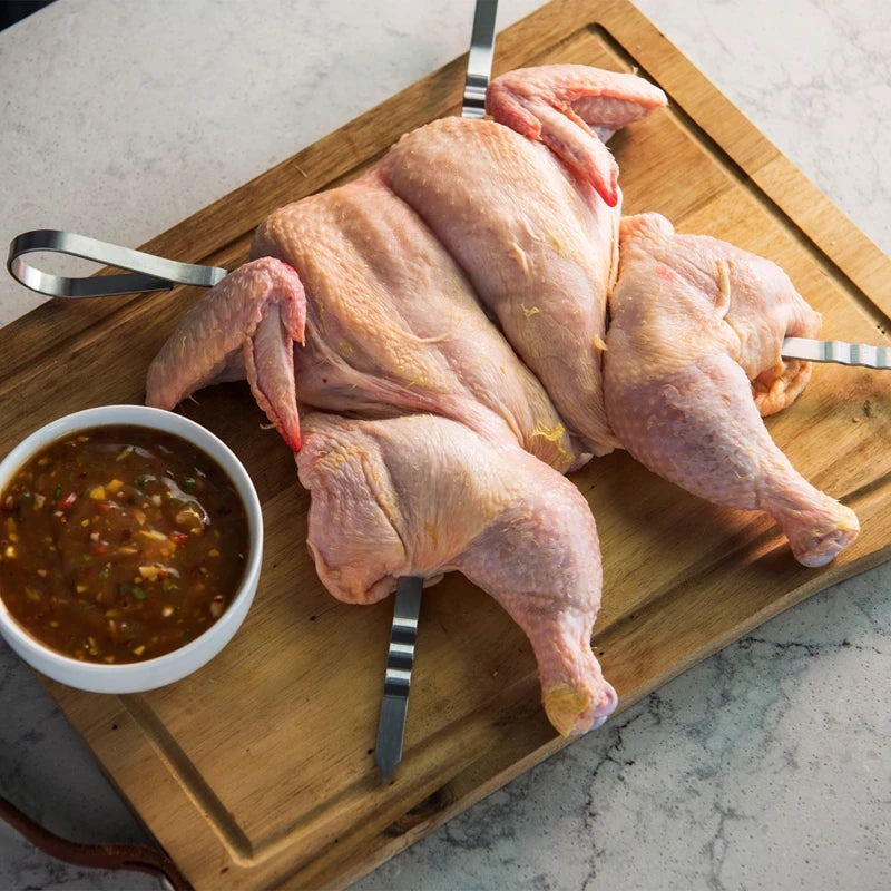 En rå kylling med Napoleon Shashlik-grillspyd hviler på et skjærebrett av tre, ved siden av en bolle med saus.