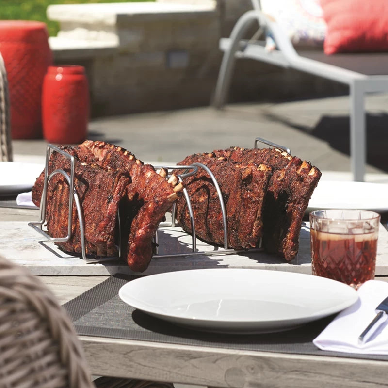 På et Napoleon Steke- og kyllingstativ på et utendørsbord ligger to ribbeholdere med tallerken, serviett og drikke, og i bakgrunnen skimtes utemøblene.