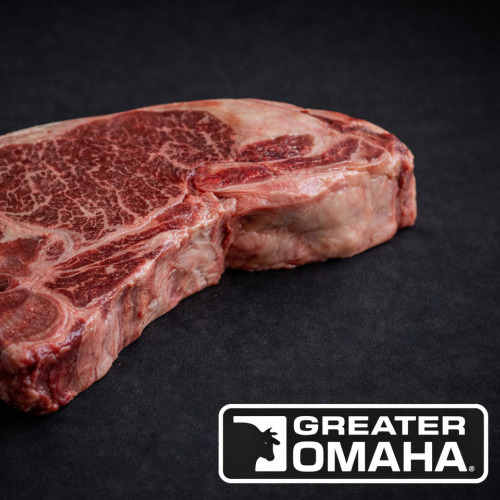 En rå, marmorert Storfe T-Bone Greater Omaha 500 g biff hviler på en svart overflate, med Greater Omaha-logoen nederst i høyre hjørne.