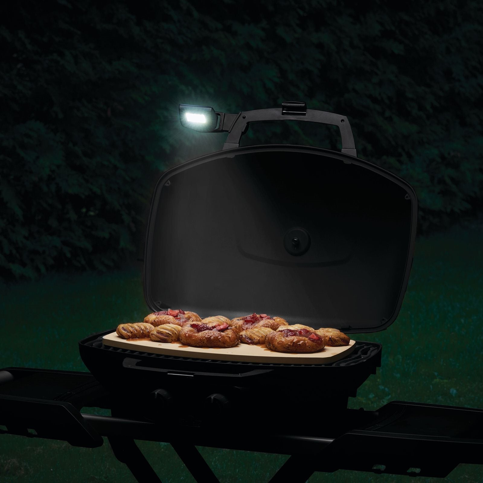 En åpen TravelQ-grill med Napoleon LED-lys til TravelQ over hodet lyser opp flere bakverk på et brett, plassert utendørs i mørket med trær i bakgrunnen.