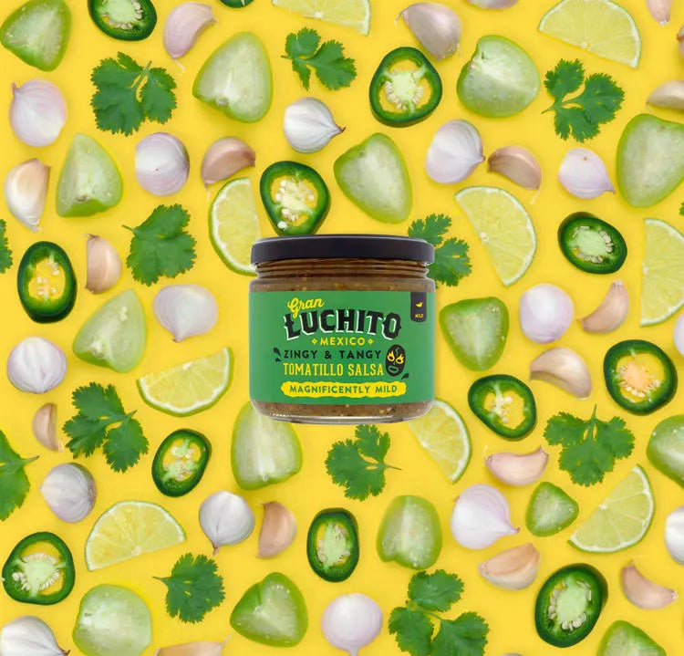 Et glass Gran Luchito Tomatillo Salsa 330 ml står på en knallgul bakgrunn, omgitt av jalapeño-skiver, tomatillo, limebåter, koriander og hvitløk - noe som skaper en livlig scene for denne meksikanske salsaen med kraftige smaker.