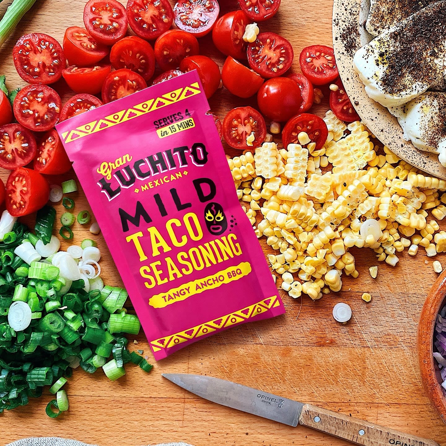 Gran Luchito Mild Taco Seasoning 28 g på en treplate, omgitt av hakket grønn løk, tomater, mais og andre friske grønnsaker.
