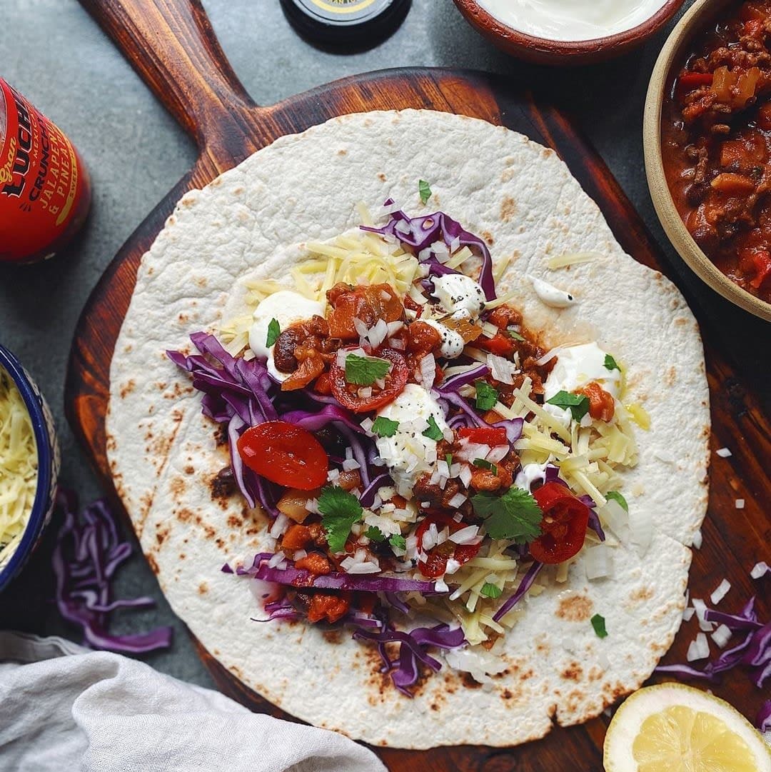 En «Gran Luchito Burrito Wrap» toppet med revet ost, rødkål, tomater, løk i terninger, urter, rømme og salsa ligger på et trebrett.