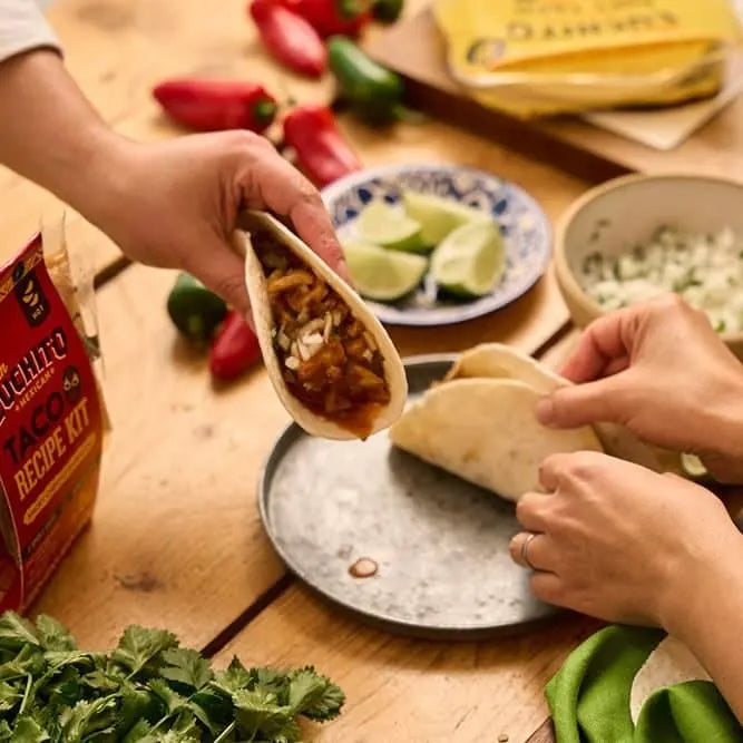 To personer som lager tacoer ved et bord med tortillas, hakket løk, lime-båter, koriander og en pakke Gran Luchito Barbacoa Recipe Kit 300 g – klare til å nyte autentiske meksikanske smaker med langtidskokt kjøtt.