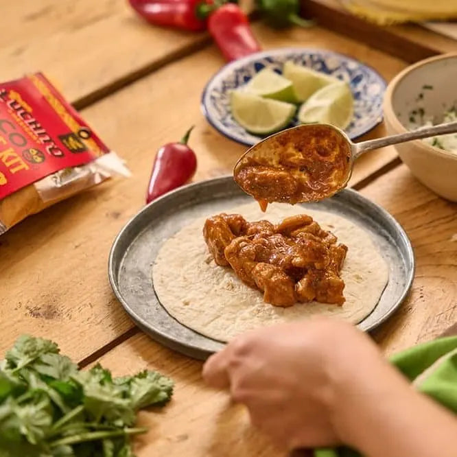 En person legger tilberedt, krydret kylling på en tortilla på en tallerken, sammen med lime-båter, urter og Gran Luchito Barbacoa Recipe Kit 300 g – som byr på autentiske meksikanske smaker – samt chipotle ved siden av på et trebord.