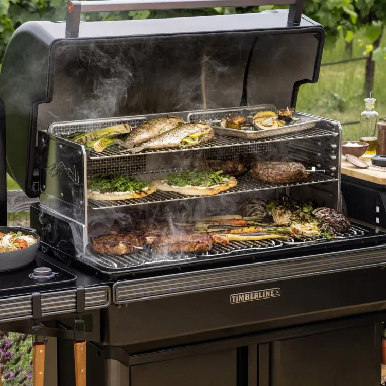 Fyr opp Traeger Timberline XL pelletsgrill, fylt med fisk, grønnsaker, skalldyr og biff på flere stativer. Nyt smakfull røyk og utendørs matlaging omgitt av frodig grønt.