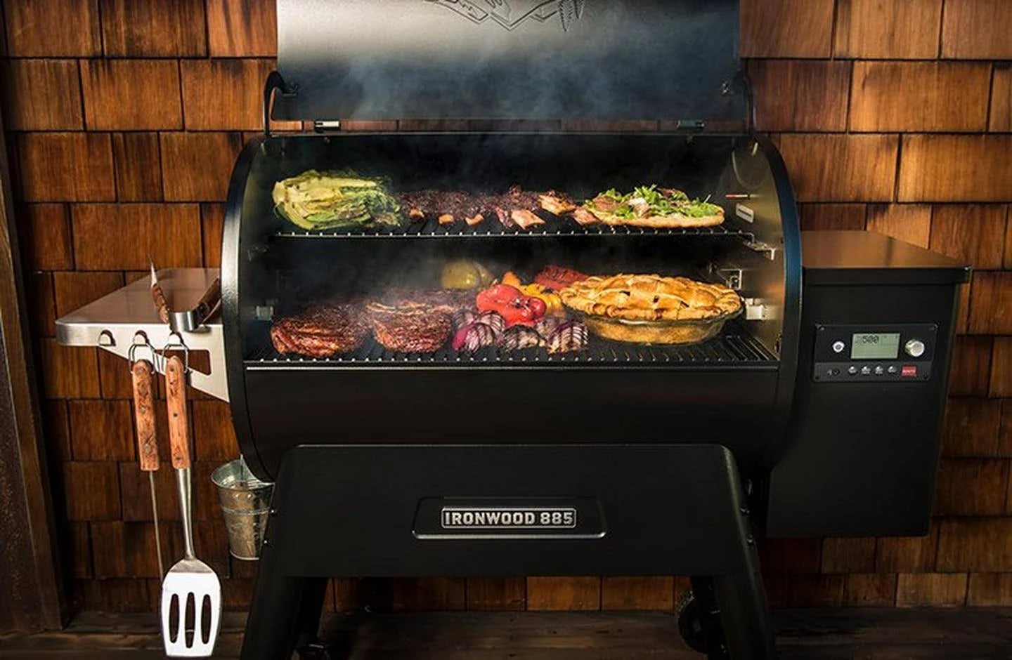 Traeger Ironwood D2 885 pelletsgrill er fylt med grønnsaker, kjøtt og en pai, mens grillredskapene henger på siden og røyken stiger opp fra innsiden.