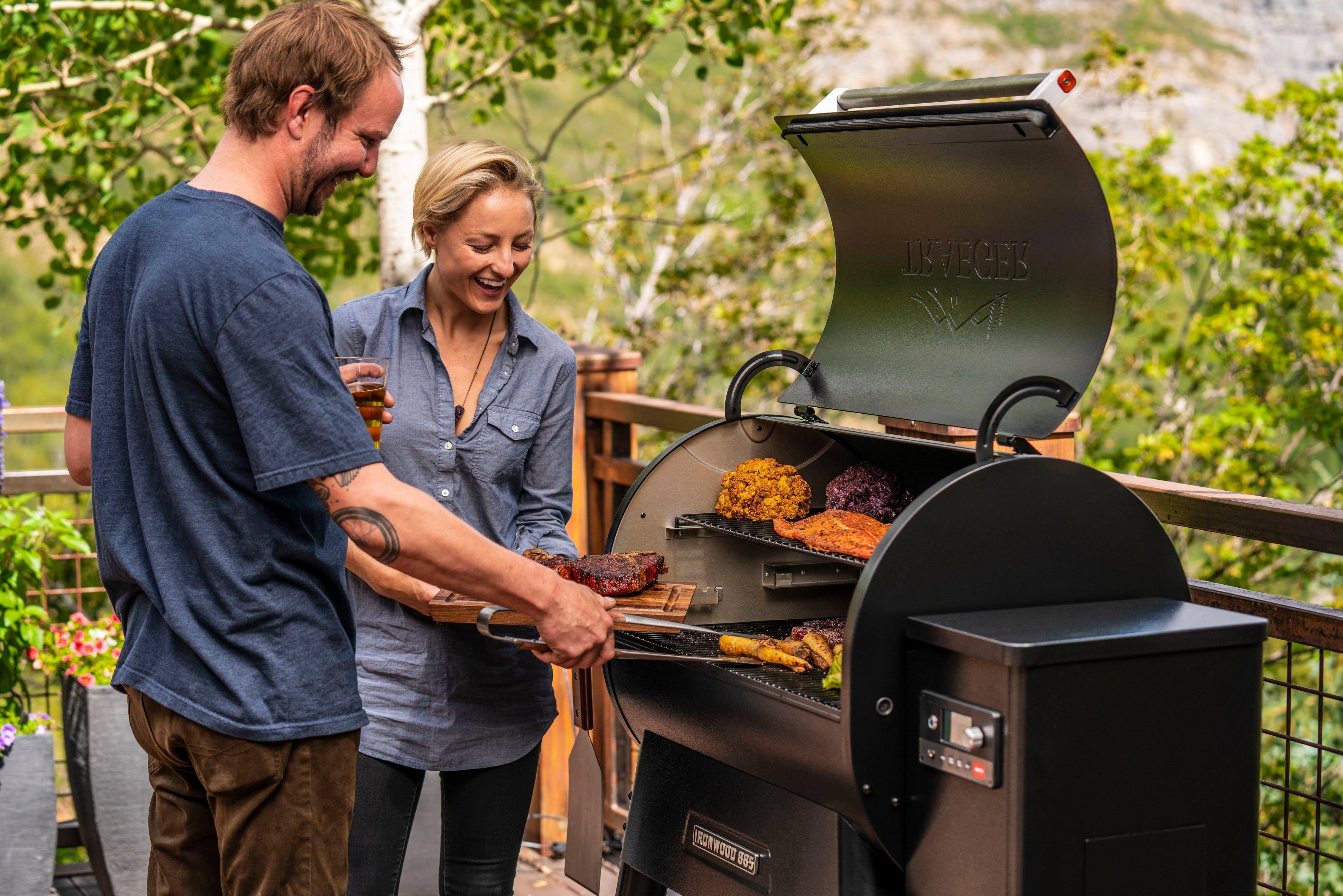 To personer står på en terrasse og bruker pelletsgrillen Traeger Ironwood D2 885. De utnytter WiFIRE-teknologien til å tilberede kjøtt og grønnsaker utendørs på en solrik dag.