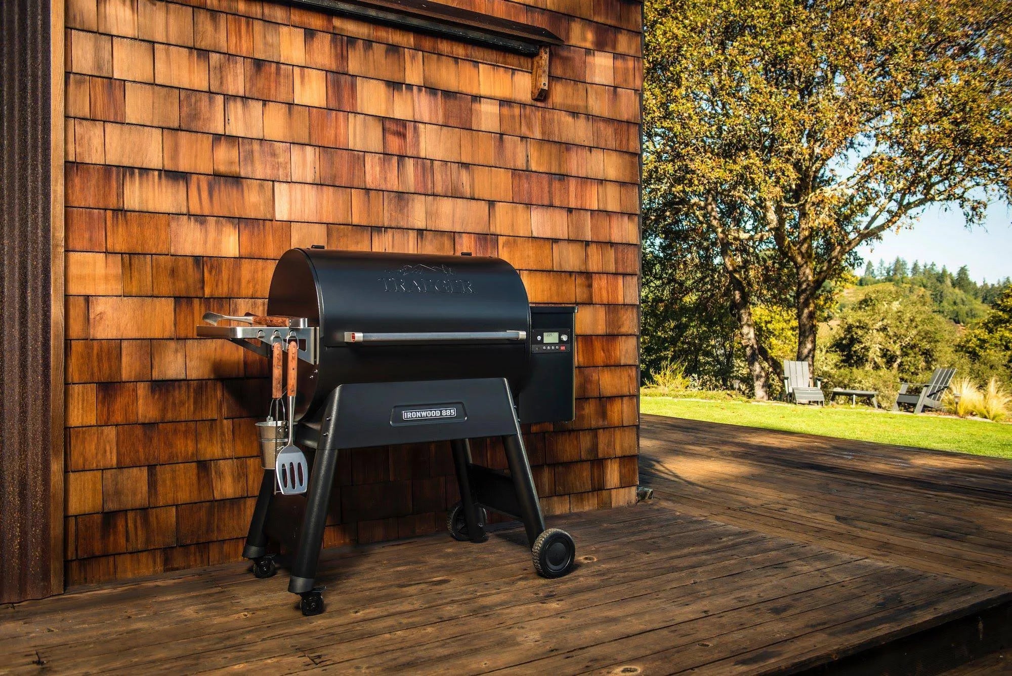 En svart Traeger Ironwood D2 885 pelletsgrill står på et tredekke ved siden av et hus med takshingel, med trær og utendørsstoler i nærheten. WiFIRE-teknologien gjør at du kan styre grillen fra hvor som helst.