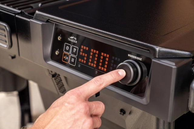 En hånd justerer temperaturknappen på Traeger Woodridge Pelletsgrill (Forhåndsbestilling) med digitalt display og WiFIRE-teknologi for presis grilling.
