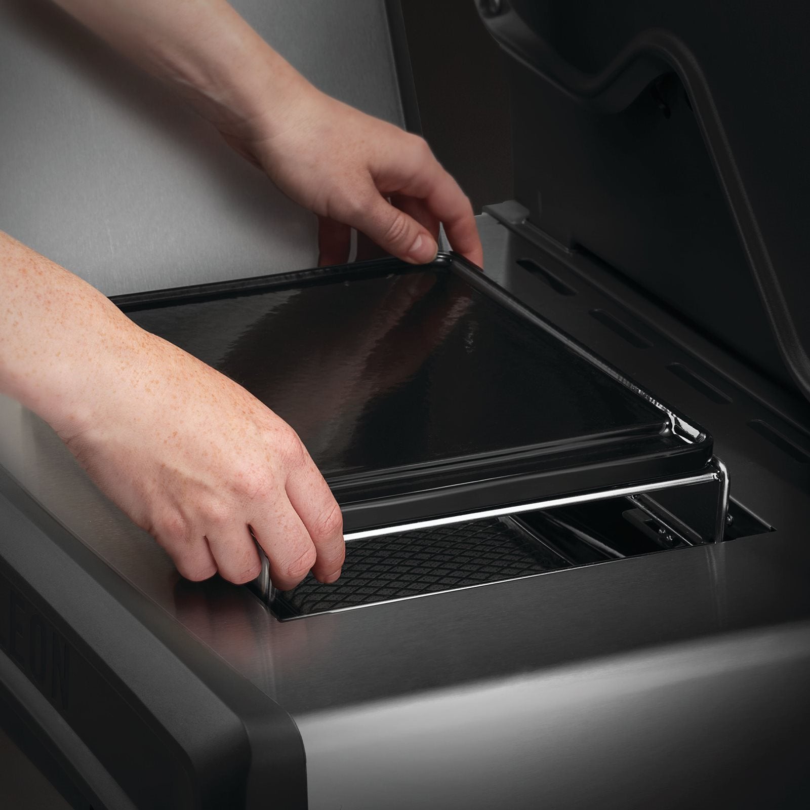 En person setter inn eller tar ut en Napoleon Stekeplate til SIZZLE ZONE, liten fra sporet på innsiden av en SIZZLE ZONE i en grill i rustfritt stål eller et kjøkkenapparat.