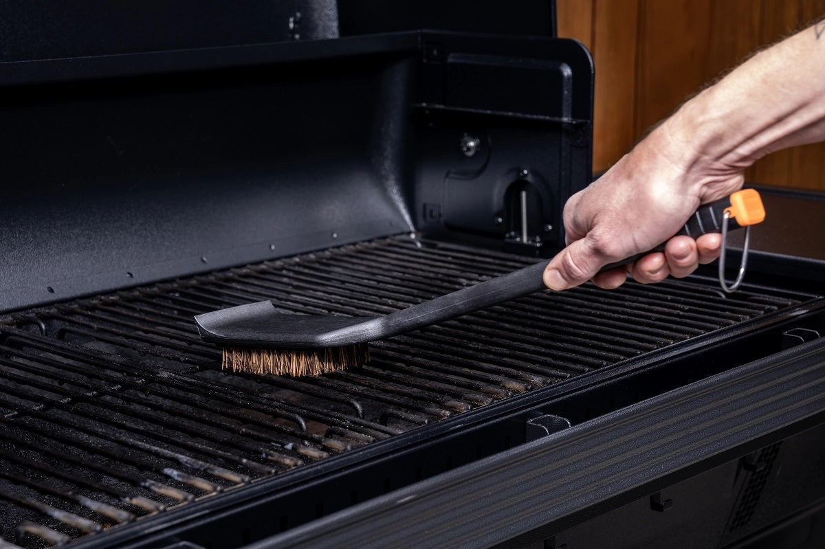 En person som rengjør en grillrist med Traeger Hot n' Cold Grill Cleaning Brush, med naturlige palmyrafibre, inne i en åpen grill for effektiv og miljøvennlig rengjøring av grillen.