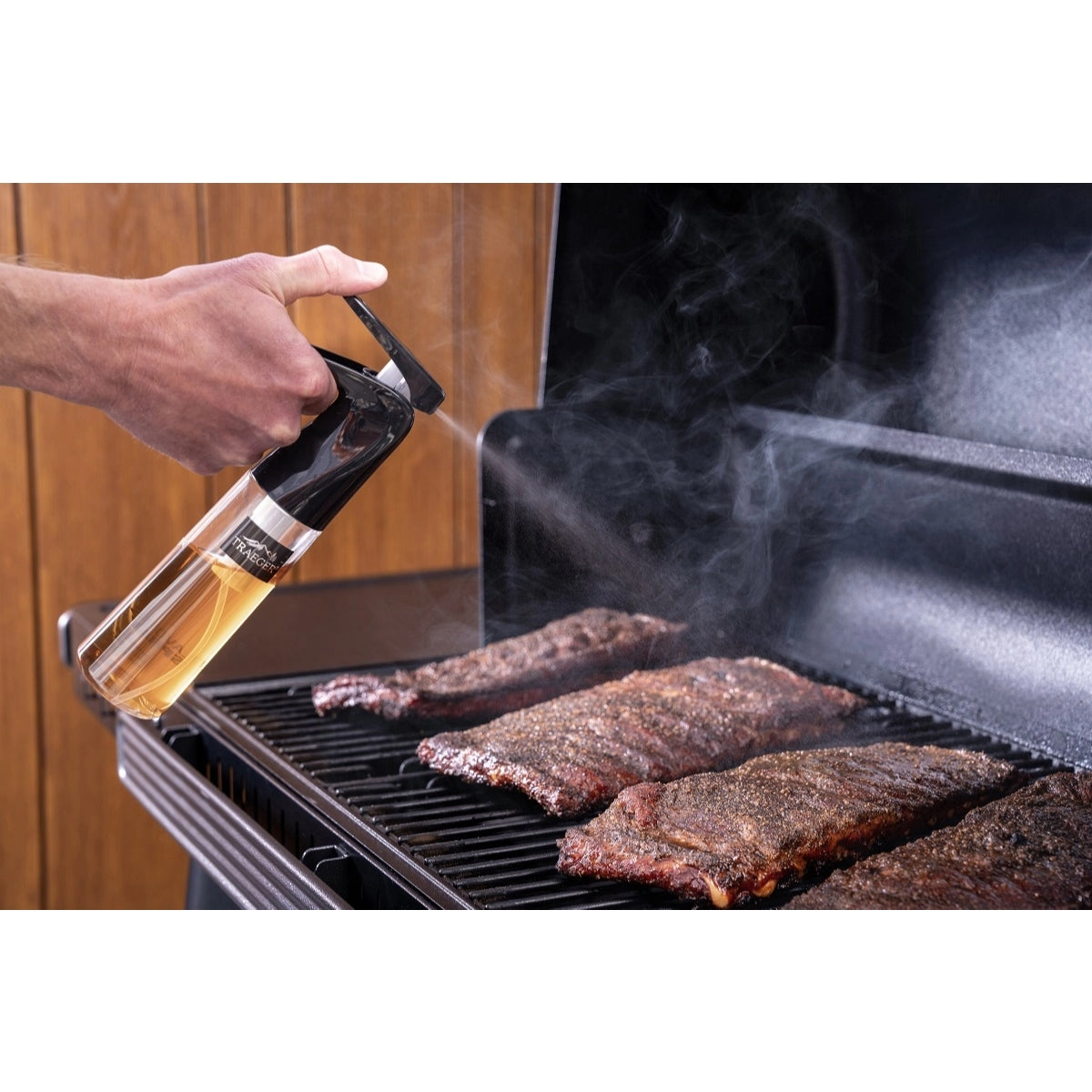 En hånd bruker Traeger BBQ Spritzer til å tåke ribbe som steker på grillen, mens røyken stiger opp fra kjøttet - et perfekt eksempel på Traeger grilltilbehør.
