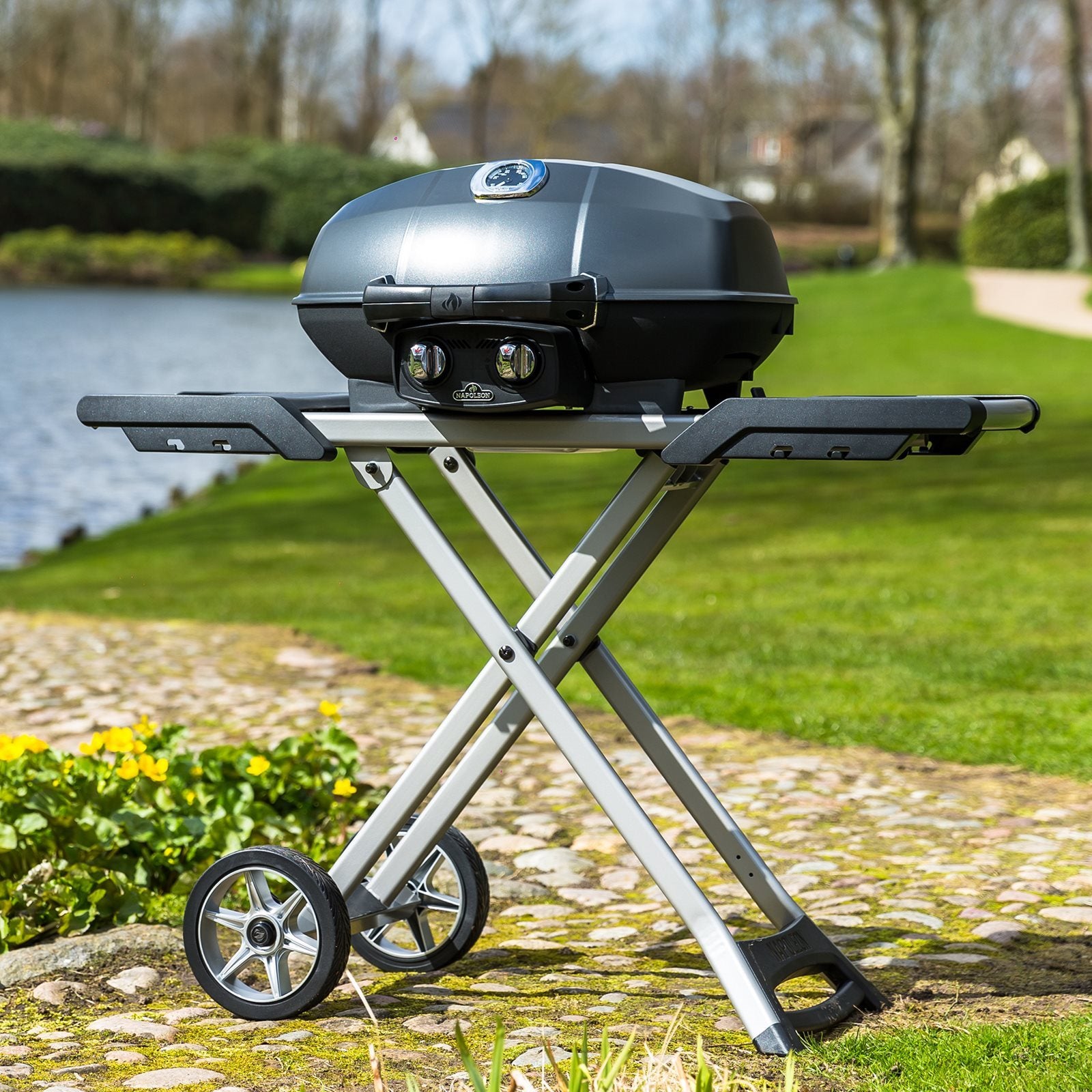 Napoleon TravelQ PRO 285X Gassgrill er en kompakt, bærbar grill med lukket lokk på et sammenleggbart stativ med hjul, plassert på en steinsti ved en dam og gule blomster i et utendørs miljø.