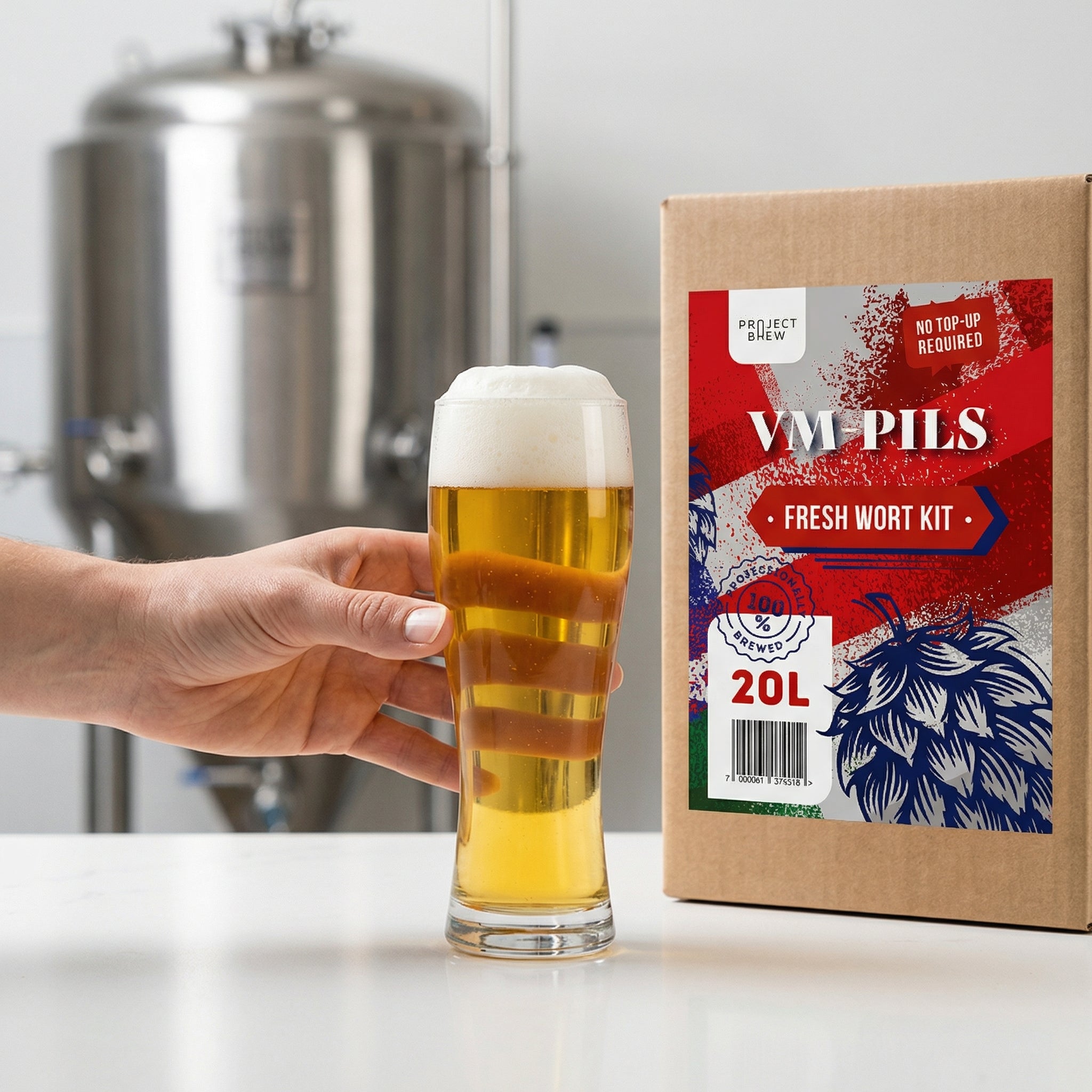 En hånd holder et høyt glass lyst øl med skum ved siden av en VM Pils - 20L Fresh Wort Kit-boks, ideell for hjemmebrygging, med bryggeriutstyr i bakgrunnen.