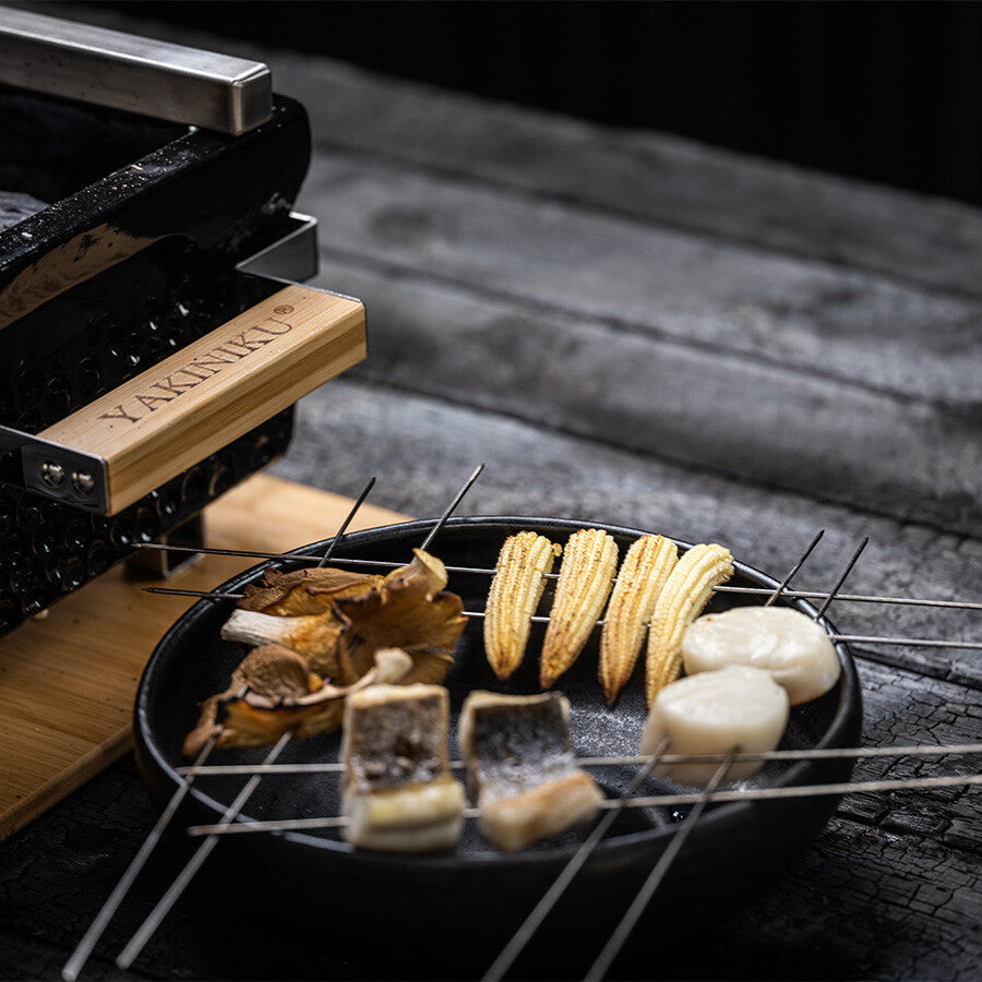 På en svart tallerken ligger YAKINIKU BBQ Spyd i Rustfritt Stål 1,0 mm 20stk med fisk, babymais, kamskjell og sopp ved siden av en Kamado yakiniku-grill på en treflate.