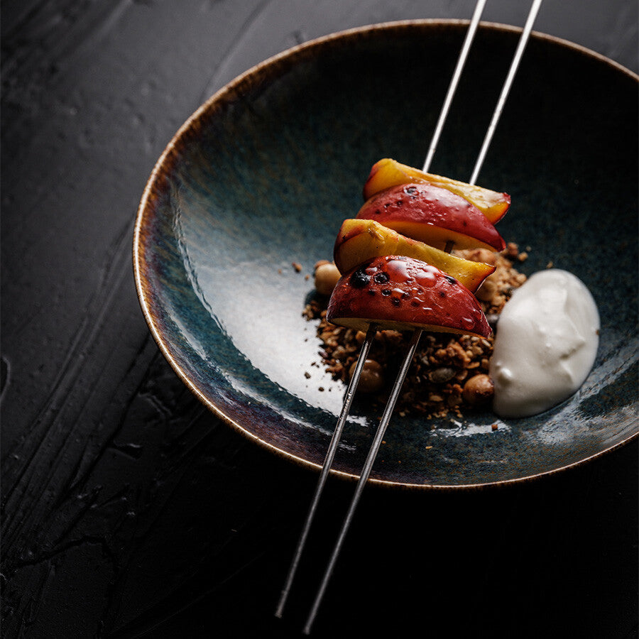 Blå keramikkskål med fruktbiter på YAKINIKU BBQ Spyd i Rustfritt Stål 2,5 mm 20stk, granola og en klatt hvit krem, satt på en mørk strukturert overflate.