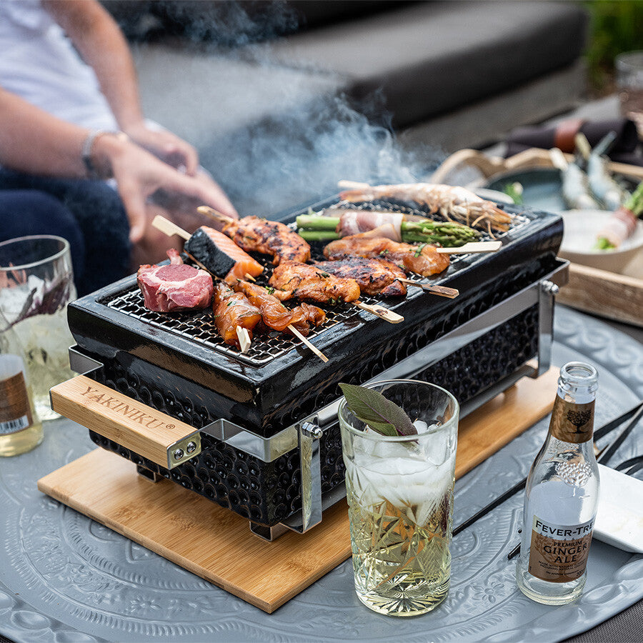 YAKINIKU Rectangular Shichirin | Konro & Yakitori Grill syder av ulike kjøtt- og grønnsaksspyd, omgitt av drikke og tilbehør - perfekt for en utendørs festmåltid.