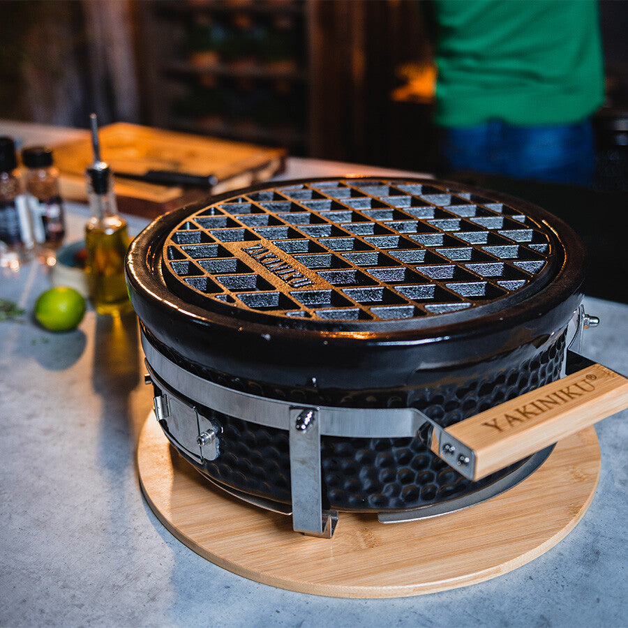 YAKINIKU Shichirin Støpejernsrist Rund, Medium, en svart bærbar grill med støpejernsrist, står på en trefot på benkeplaten, med krydder og redskaper i nærheten.